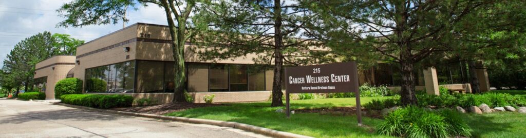 A single-story brick building with large windows surrounded by green trees and shrubs. A sign in front reads “Garden Wellness Center.” The sky is partly cloudy, and the area appears well-maintained.