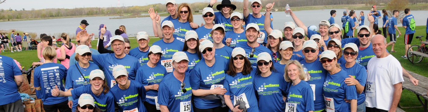 A large group of smiling runners wearing matching blue shirts and white caps pose together outdoors by a lake before a race. Some people hold race numbers; others wave or give thumbs up. More participants are in the background.
