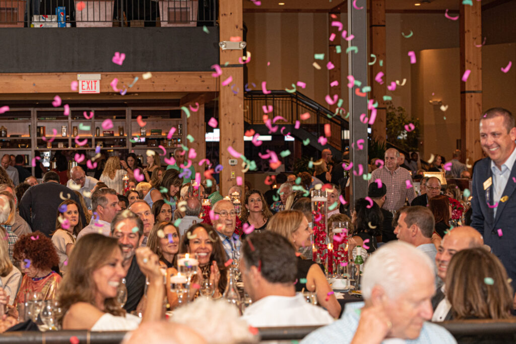 A lively crowd sits at tables in a decorated indoor venue, watching colorful confetti fall from above. People are smiling and celebrating, with candles and floral centerpieces on the tables.
