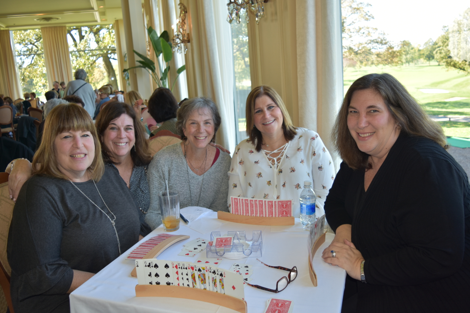 Five women are smiling at a table in a bright restaurant, playing cards together. Two card holders with visible playing cards and drinks are on the table. Large windows show trees and greenery outside.