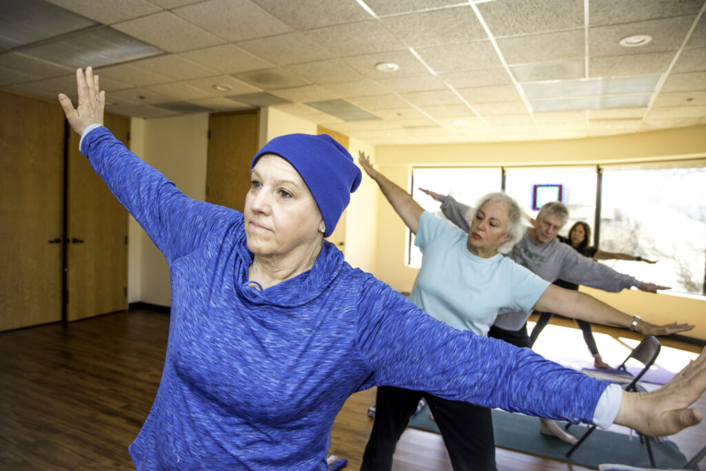 Four older adults practice yoga indoors, standing in a row with arms outstretched in a warrior pose. The group is focused, and the bright room has wood floors and large windows letting in natural light.