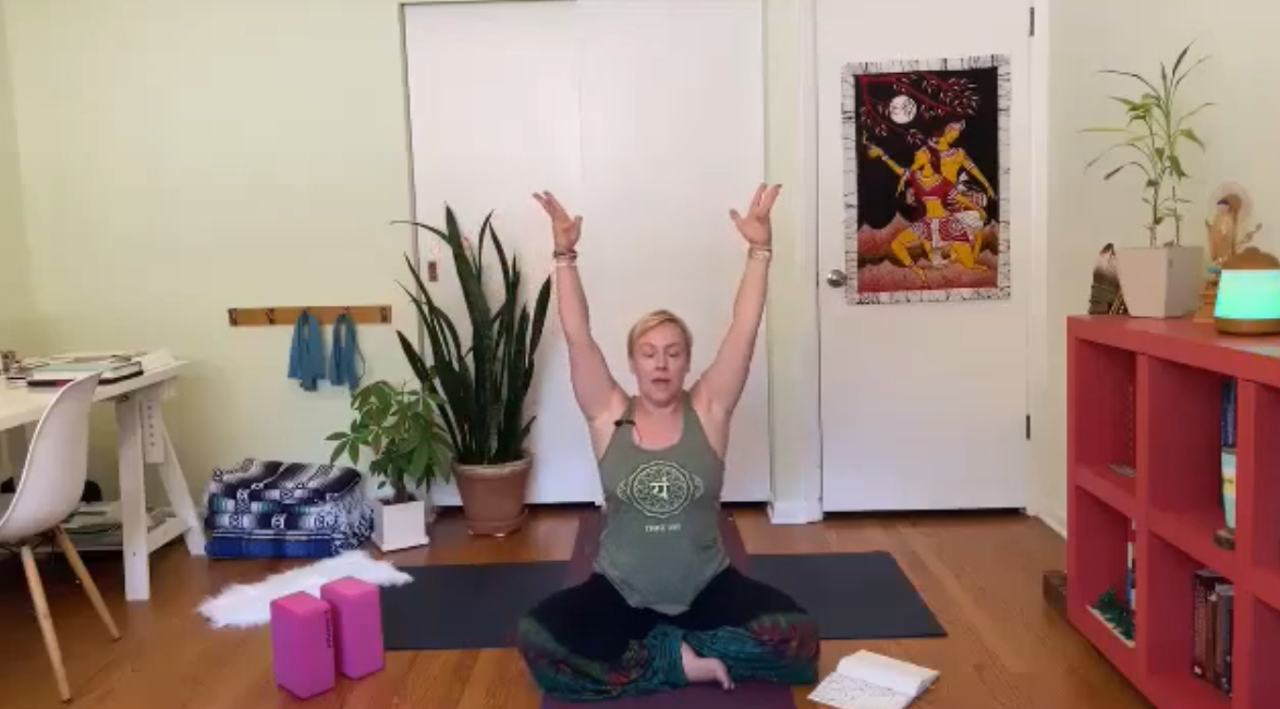 A woman sits cross-legged on a yoga mat indoors, raising both arms overhead. Yoga props, plants, and bookshelves surround her. She appears to be leading a virtual yoga or meditation session.