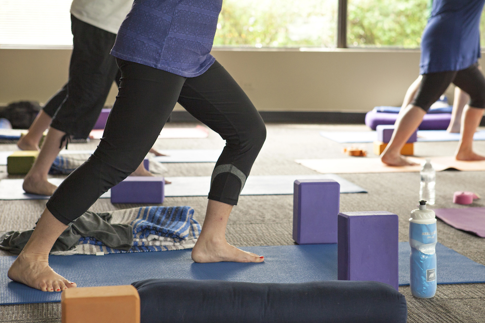 People practice yoga indoors on mats, surrounded by yoga blocks, towels, and water bottles. The focus is on their legs and feet in a standing pose, with natural light coming through large windows in the background.