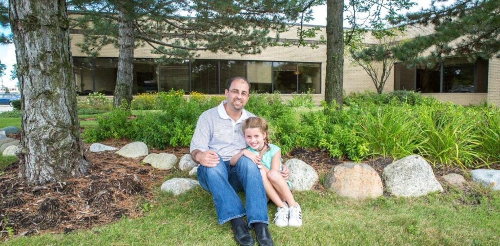 A man and a young girl sit together on grass in front of a landscaped area with rocks, bushes, and trees, smiling at the camera. A brick building with large windows is in the background.