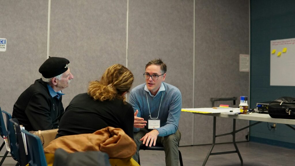 Three people are sitting and having a discussion in a conference room. One man in a blue sweater speaks while another man in a beret and a woman listen. A table with papers and supplies is in the background.