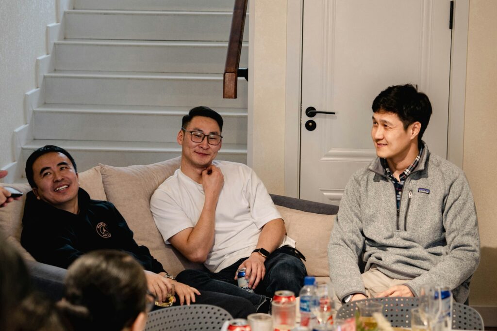 Three men sit on a couch, smiling and conversing in a cozy indoor setting. A staircase and white door are visible in the background, with drinks and snacks on the table in front of them.