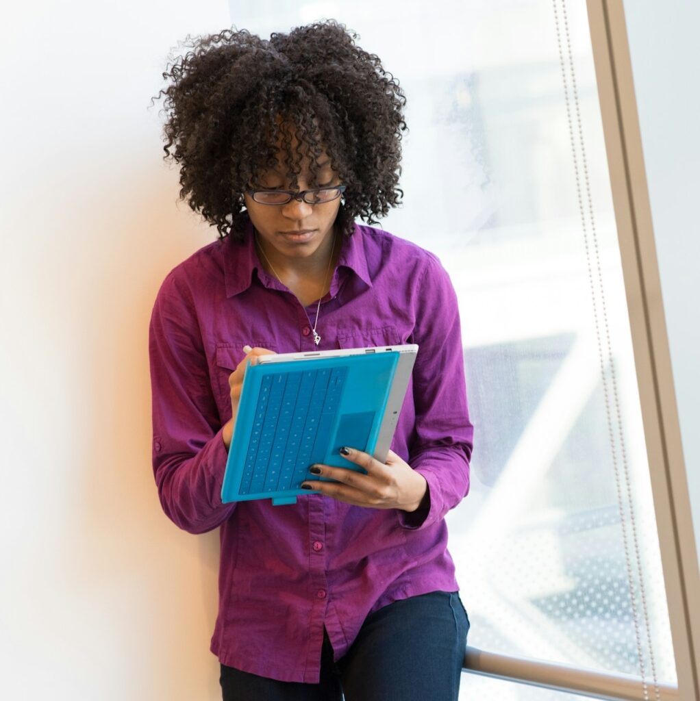 A person with curly hair and glasses, wearing a purple shirt, stands by a window while writing on a blue tablet with a stylus.