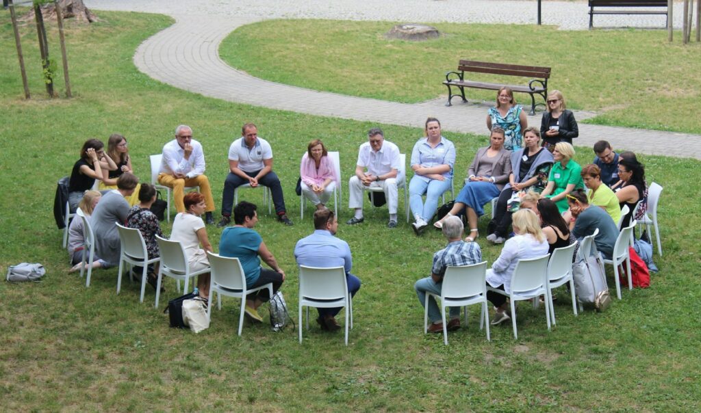 A group of people sit in a circle on white chairs outside on grass, engaged in conversation. The meeting takes place in a park-like setting with paths, benches, and trees in the background.
