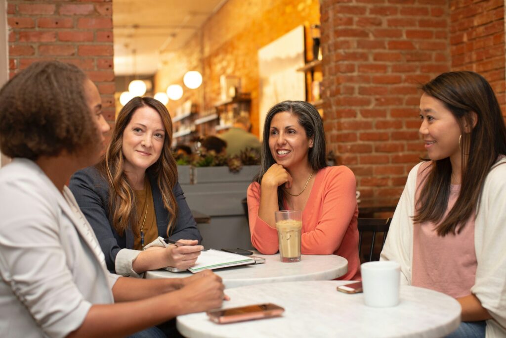 Four women sit around a table in a cozy café, engaged in a friendly conversation. One woman holds a notepad, another has an iced coffee, and a third has a white mug. They are all smiling and appear to be enjoying their discussion.