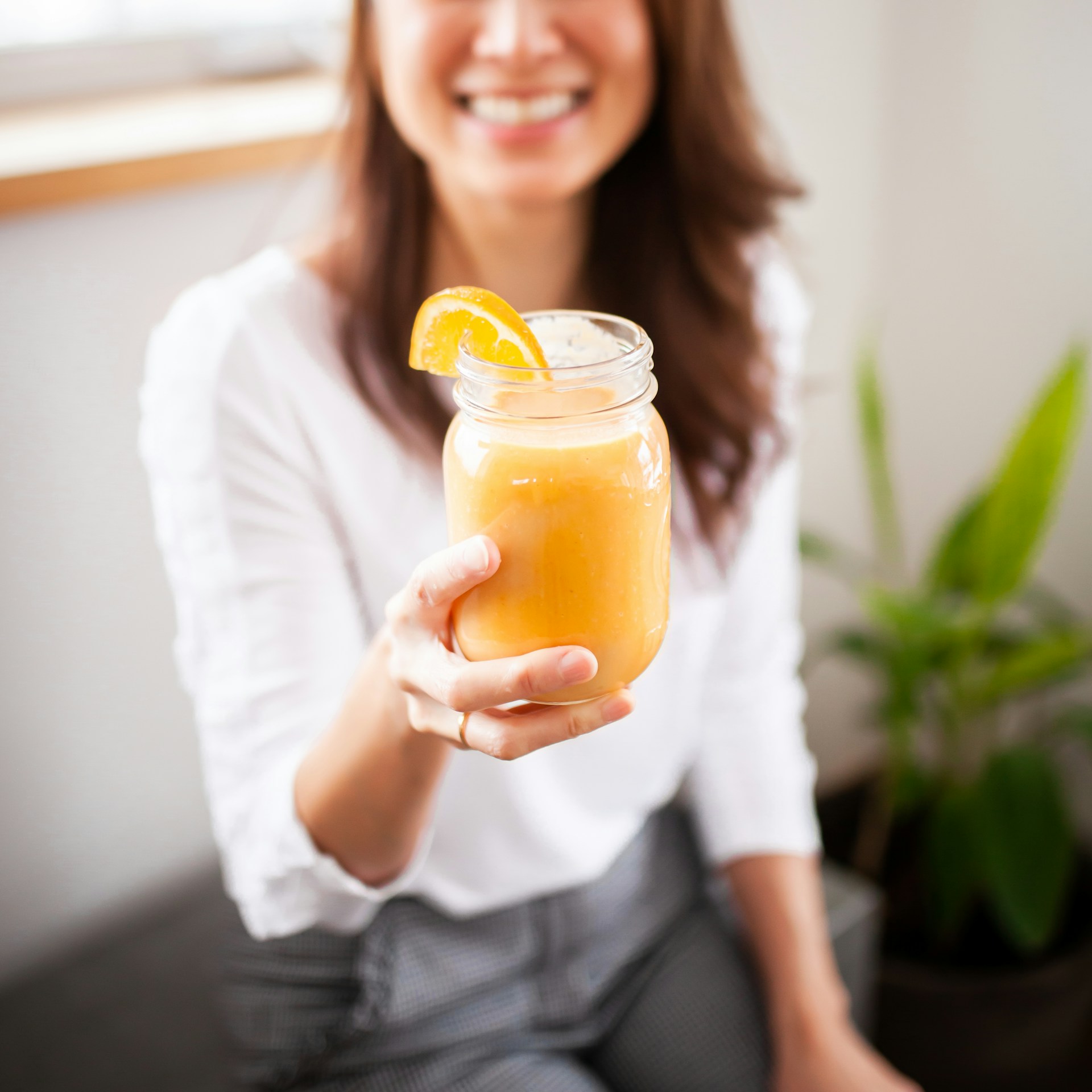 A smiling woman in a white top holds a mason jar of orange smoothie garnished with an orange slice, offering it toward the camera. A green plant is blurred in the background.