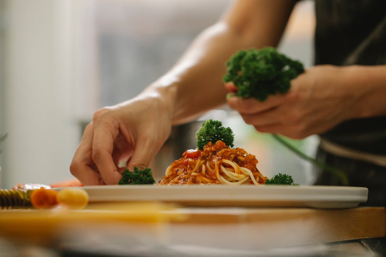 A person garnishing a plate of spaghetti with parsley, using their hands to carefully place fresh green herbs around the pasta topped with tomato sauce.