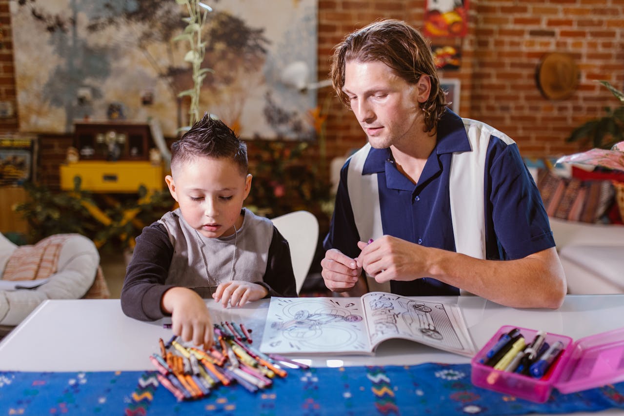A man and a young boy sit at a table coloring in a book with crayons. The boy is reaching for crayons while the man watches and offers guidance. The setting appears to be a cozy, art-filled room.