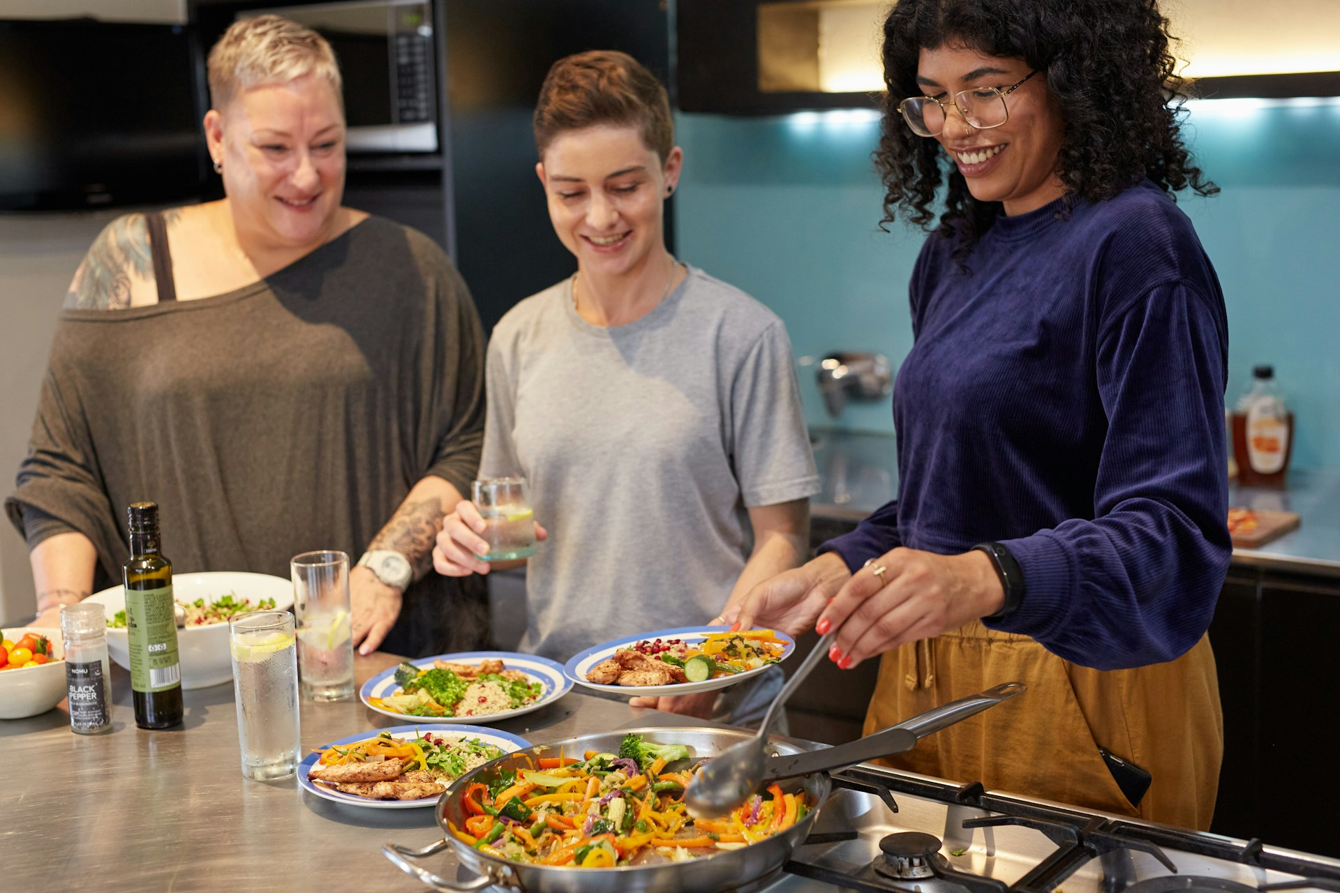 Three people stand in a kitchen, smiling and preparing or serving colorful dishes from pans on the stove, with plates of food and drinks in front of them.