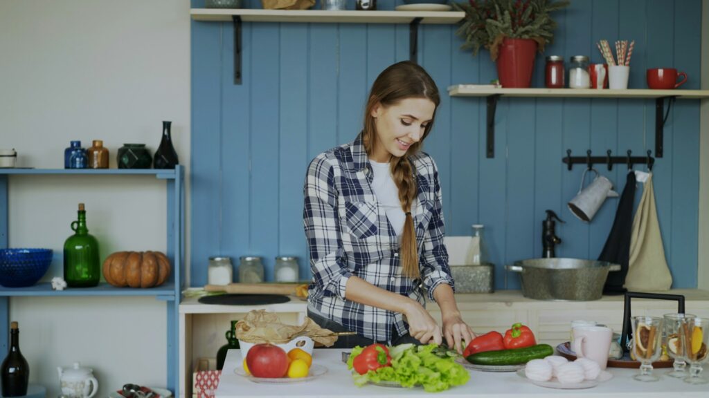 A woman in a plaid shirt smiles while chopping vegetables in a bright kitchen. Fresh produce like lettuce, tomatoes, and cucumbers are on the counter, with shelves and kitchenware visible in the background.
