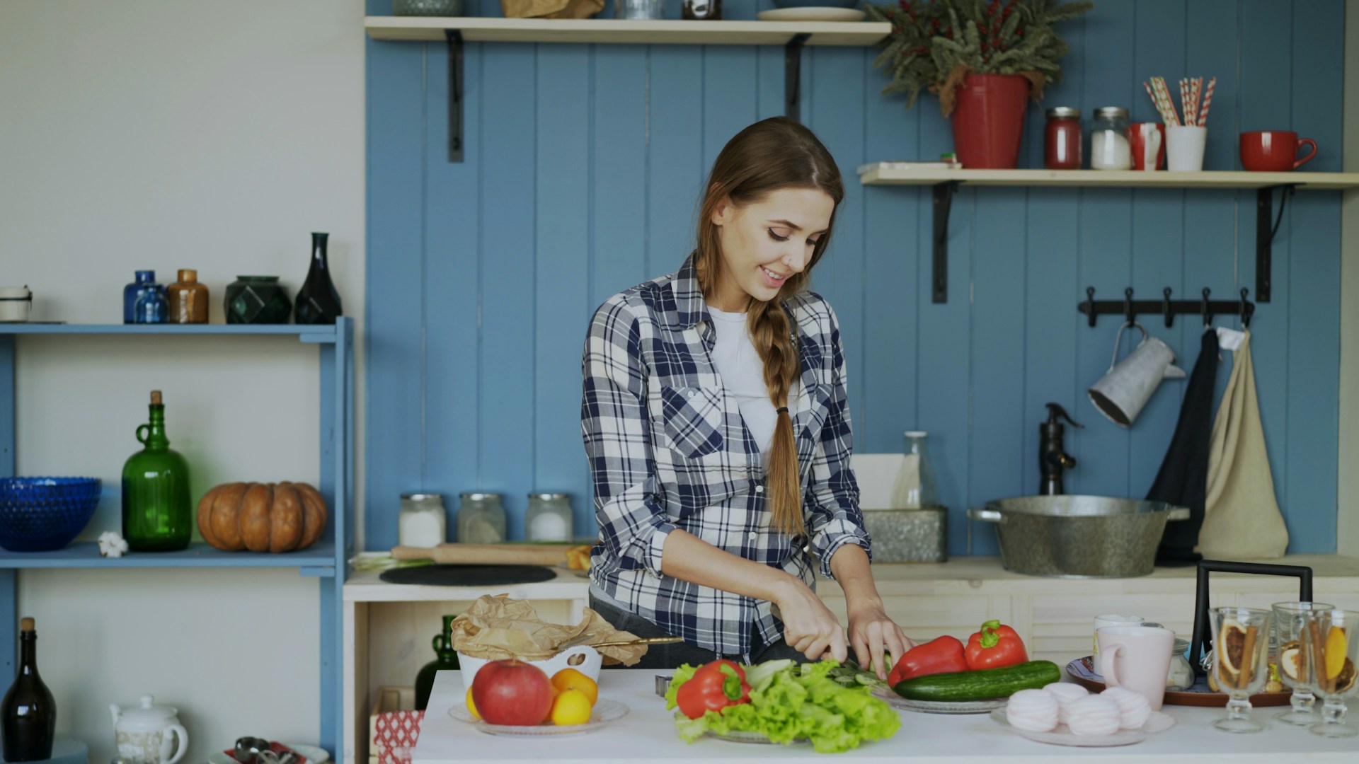 A woman in a plaid shirt smiles while chopping vegetables in a bright kitchen. Fresh produce like lettuce, tomatoes, and cucumbers are on the counter, with shelves and kitchenware visible in the background.