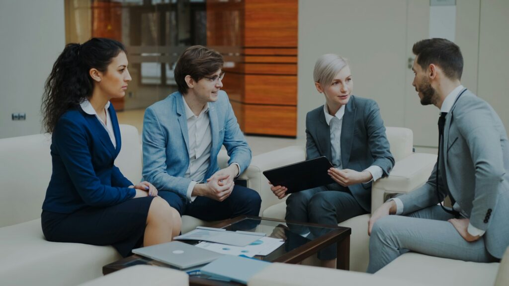 Four business professionals, two women and two men, sit on couches in an office lounge, having a discussion. One woman holds a tablet while documents and a laptop are on the coffee table in front of them.