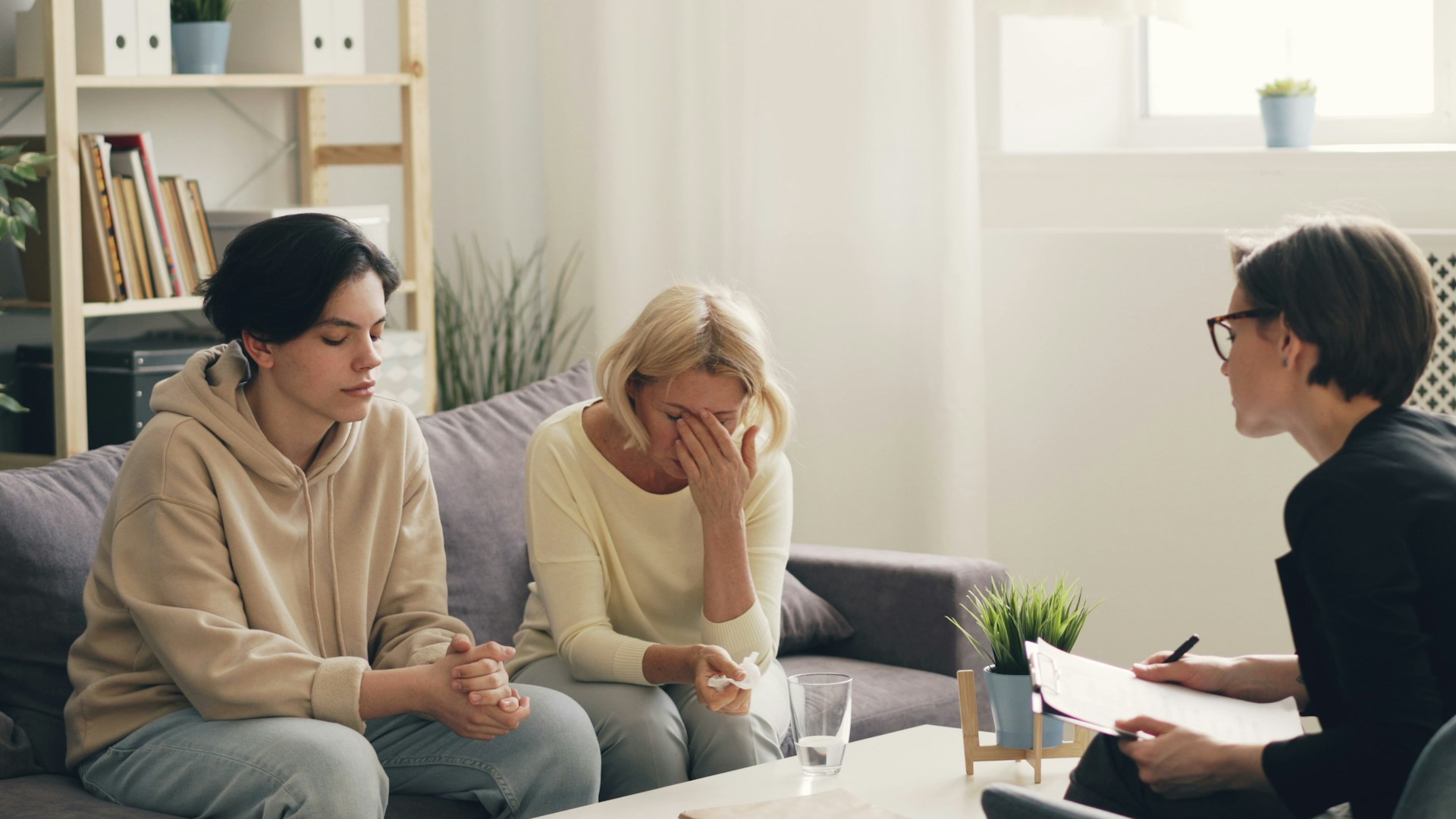 Two women sit on a couch, one with her head in her hand looking distressed and the other looking down pensively, while a third person, possibly a therapist, sits across from them holding a clipboard.