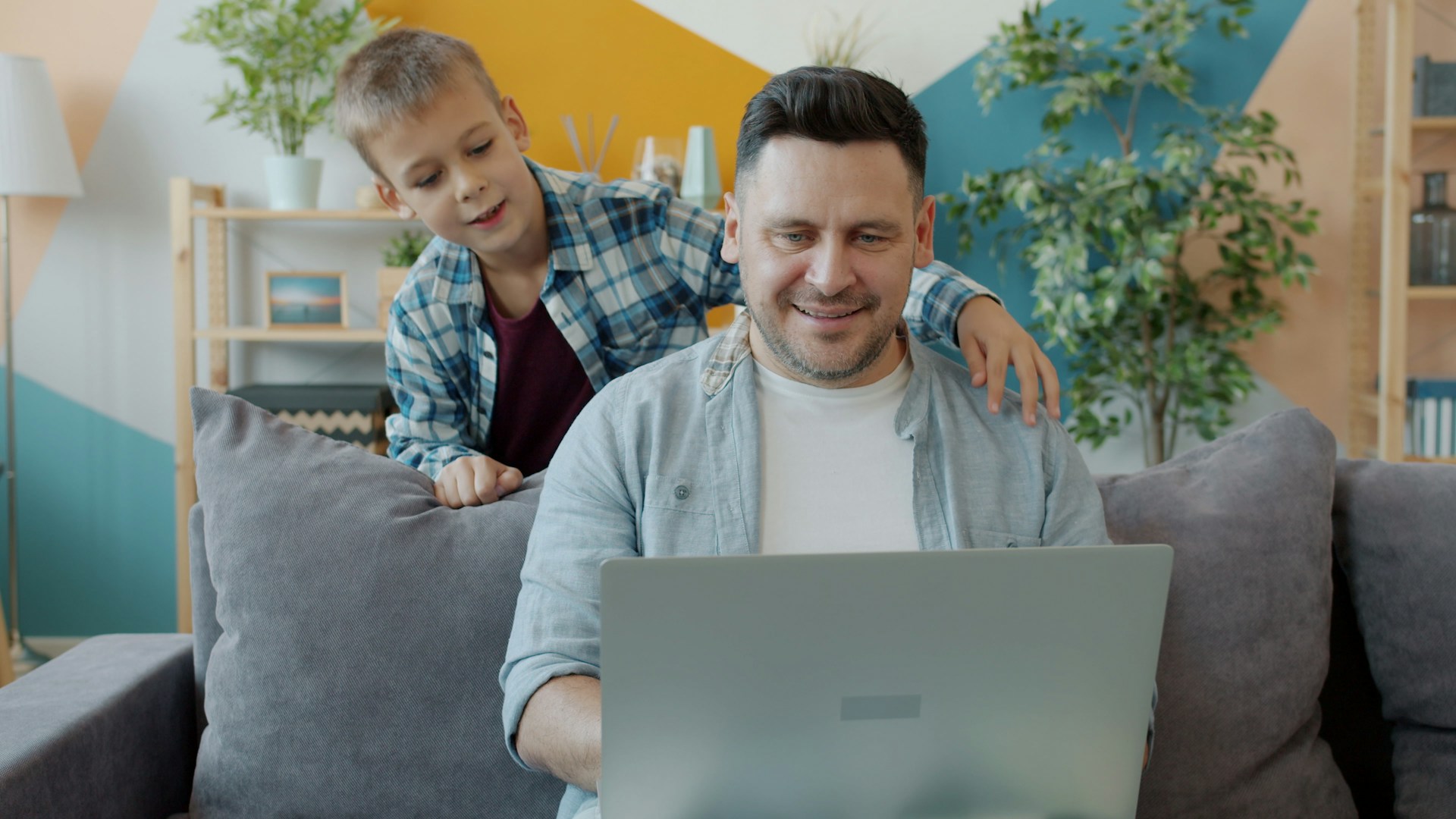 A man sits on a gray couch using a laptop while a young boy stands behind him with an arm on his shoulder. Both appear happy and relaxed in a bright, modern living room with plants and colorful walls.