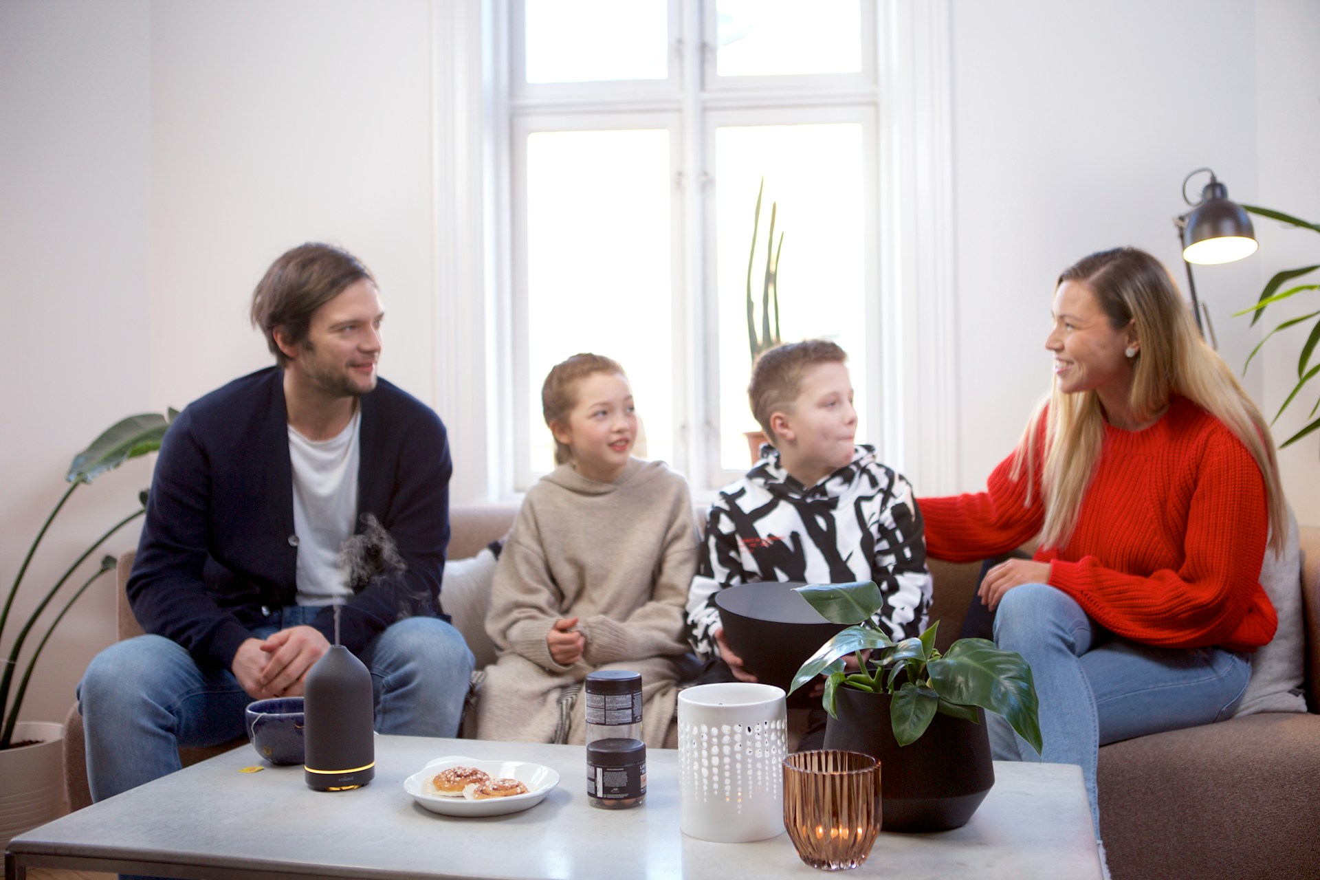 A family of four sits on a living room couch, smiling and talking. Two children are in the middle, with an adult man on the left and an adult woman on the right. A coffee table with snacks, plants, and decor is in front of them.