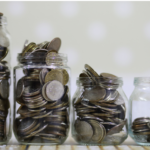 Four glass jars containing varying amounts of coins are lined up in a row from fullest to least full, with the last jar only partially filled. The background is soft and out of focus.