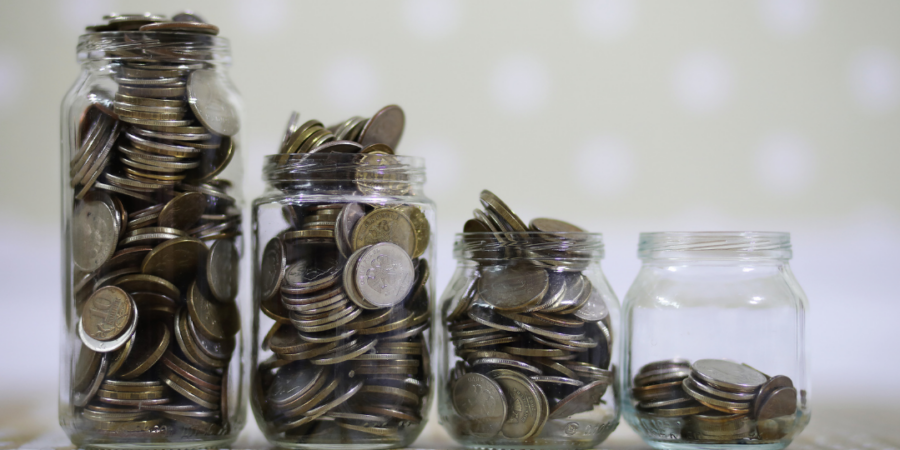 Four glass jars containing varying amounts of coins are lined up in a row from fullest to least full, with the last jar only partially filled. The background is soft and out of focus.