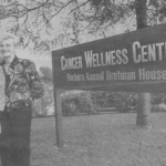 Two older women stand and smile beside a large sign that reads "Cancer Wellness Center, Barbara Kassel Brotman House" outside on a grassy area near a parking lot and trees.
