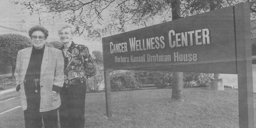 Two older women stand and smile beside a large sign that reads "Cancer Wellness Center, Barbara Kassel Brotman House" outside on a grassy area near a parking lot and trees.