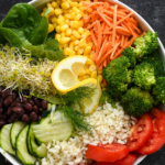A bowl filled with fresh vegetables including spinach, corn, shredded carrot, broccoli, tomato, cucumber, black beans, alfalfa sprouts, and cauliflower, garnished with lemon slices and dill, on a dark surface.