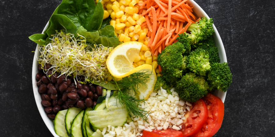 A bowl filled with fresh vegetables including spinach, corn, shredded carrot, broccoli, tomato, cucumber, black beans, alfalfa sprouts, and cauliflower, garnished with lemon slices and dill, on a dark surface.
