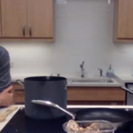 Two women are in a kitchen. One is sitting at a counter with ingredients and cookware, speaking, while the other sits in the background wearing an apron. Various cooking supplies and vegetables are visible on the counter.