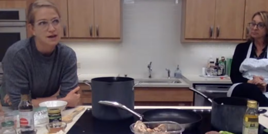 Two women are in a kitchen. One is sitting at a counter with ingredients and cookware, speaking, while the other sits in the background wearing an apron. Various cooking supplies and vegetables are visible on the counter.