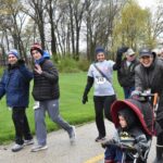 A group of people bundled in jackets and hats walk in a park during a race event. Some wave and smile at the camera, while a child in a red stroller dressed as Batman rides along. Trees with early spring leaves are in the background.