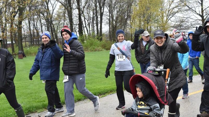 A group of people bundled in jackets and hats walk in a park during a race event. Some wave and smile at the camera, while a child in a red stroller dressed as Batman rides along. Trees with early spring leaves are in the background.