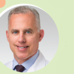 A middle-aged man with short gray hair, wearing a white lab coat, light blue shirt, and maroon tie, smiling. The background is light green with pink and orange dots.