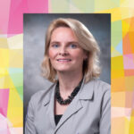 A woman with blonde hair wearing a light gray blazer and black beaded necklace poses against a gray background. The portrait is centered on a colorful geometric pattern backdrop.