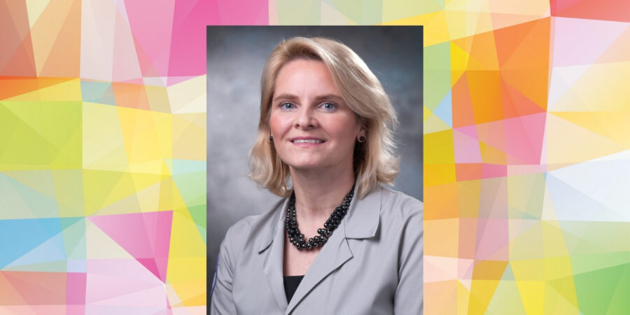 A woman with blonde hair wearing a light gray blazer and black beaded necklace poses against a gray background. The portrait is centered on a colorful geometric pattern backdrop.
