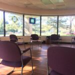 A sunlit room with large windows, stained glass panels, and nine empty purple chairs arranged in a circle on a wooden floor. Trees and parked cars are visible outside.