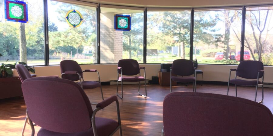 A sunlit room with large windows, stained glass panels, and nine empty purple chairs arranged in a circle on a wooden floor. Trees and parked cars are visible outside.