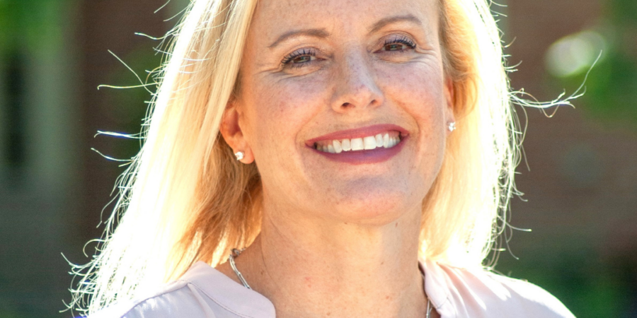 A smiling woman with long blonde hair stands outdoors in sunlight, wearing a light pink blouse and a silver necklace. The background is blurred with greenery and a brick wall.