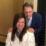 A smiling man stands behind a smiling woman who is seated. The man has his hands on the woman’s shoulders. Both are looking at the camera. The woman wears a white top with embroidery and the man wears a blue blazer.