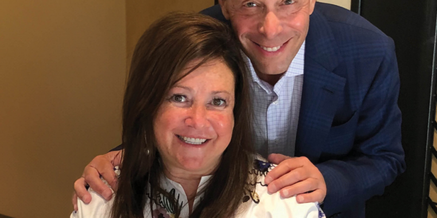 A smiling man stands behind a smiling woman who is seated. The man has his hands on the woman’s shoulders. Both are looking at the camera. The woman wears a white top with embroidery and the man wears a blue blazer.