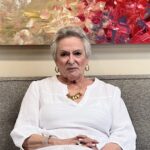 An older woman with short gray hair, gold hoop earrings, and a gold necklace sits on a gray couch wearing a white blouse. A colorful abstract painting hangs on the wall behind her.