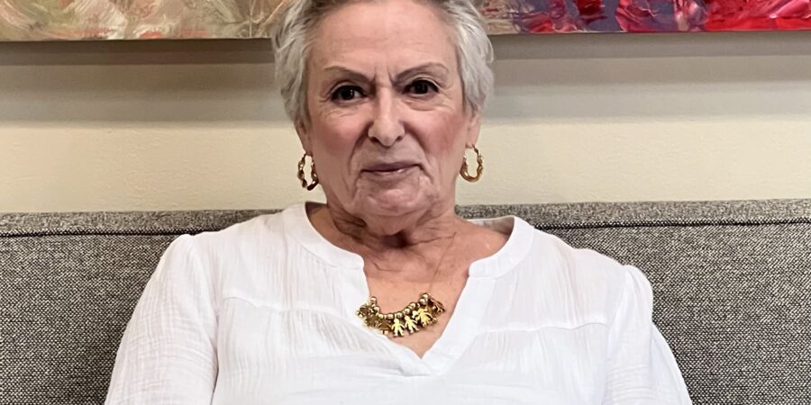 An older woman with short gray hair, gold hoop earrings, and a gold necklace sits on a gray couch wearing a white blouse. A colorful abstract painting hangs on the wall behind her.