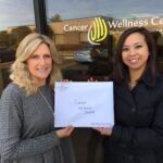 Two women stand outside a building with a “Cancer Wellness Center” sign, smiling and holding a paper that reads "Cancer Wellness Center." The reflection of a parking lot and trees is visible in the window behind them.