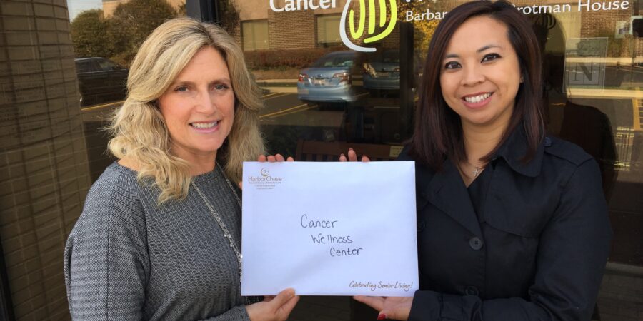 Two women stand outside a building with a “Cancer Wellness Center” sign, smiling and holding a paper that reads "Cancer Wellness Center." The reflection of a parking lot and trees is visible in the window behind them.