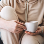 A person in beige loungewear sits with one knee up, holding a white mug with both hands, suggesting a relaxed and cozy moment. Their face is not visible.