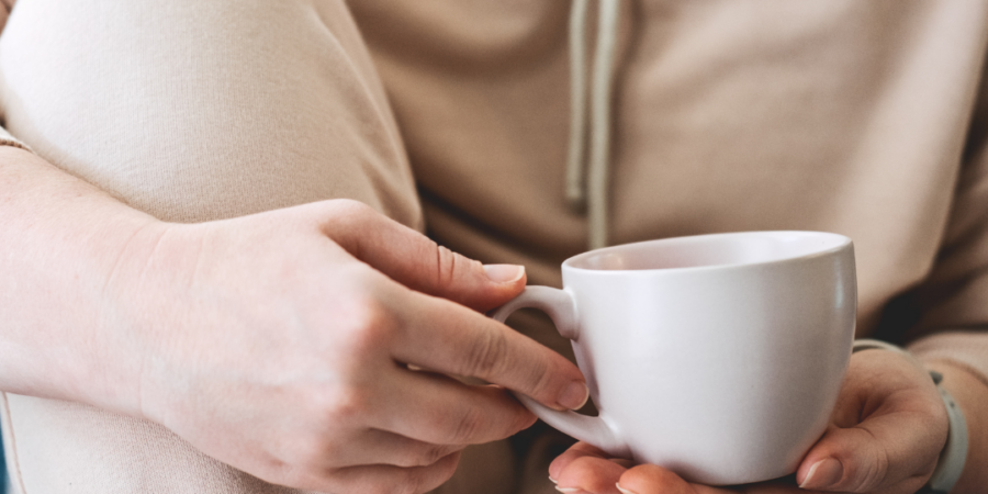 A person in beige loungewear sits with one knee up, holding a white mug with both hands, suggesting a relaxed and cozy moment. Their face is not visible.