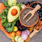 A heart-shaped platter with avocado, salmon, leafy greens, blueberries, almonds, chia seeds, tomatoes, cucumber, onion, walnuts, grapes, and various supplements on a gray background.