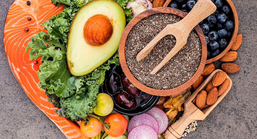 A heart-shaped platter with avocado, salmon, leafy greens, blueberries, almonds, chia seeds, tomatoes, cucumber, onion, walnuts, grapes, and various supplements on a gray background.