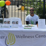 A man wearing sunglasses and a Cancer Wellness Center t-shirt sits behind an information table covered with brochures and flyers. The tablecloth reads "Cancer Wellness Center" and colorful balloons are attached to a fence behind him.